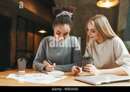 Porträt von zwei Multirassischen junge weibliche Freunde diskutieren neue Ideen und Notizen in ein Notebook, wie sie in der Cafeteria genießen eine Tasse Kaffee sitzen. Charts, Diagramme und Laptop auf dem Tisch. Stockfoto