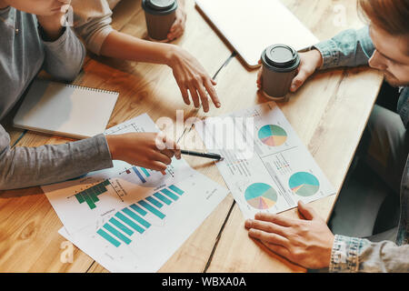 7/8 Schuß von drei Kollegen Austausch neuer Ideen miteinander, wie sie in der Cafeteria genießen eine Tasse Kaffee sitzen. Charts, Diagramme und Laptop auf dem Tisch. Horizontale erschossen. Hohen Winkel Stockfoto