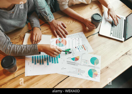 7/8 Schuß von drei Kollegen Austausch neuer Ideen miteinander, wie sie in der Cafeteria genießen eine Tasse Kaffee sitzen. Charts, Diagramme und Laptop auf dem Tisch. Horizontale erschossen. Hohen Winkel Stockfoto