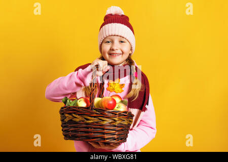 Süße kleine Mädchen in einem Schal und Hut auf einem gelben Hintergrund. Ein Kind hält einen Korb mit Äpfeln. Ernte und Herbst Konzept. Stockfoto