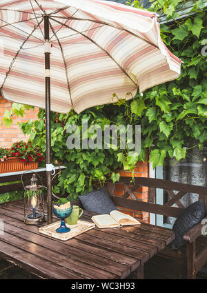 Niedlich private Balkon mit üppigen Weinreben als Dekoration und eröffnet sun Sonnenschirm für den Schatten im sonnigen Sommertag Wetter. Geöffnete Buch, moderne Stockfoto