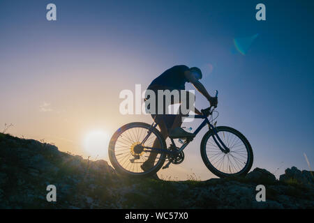 Silhouette eines passenden männlichen Mountain Biker, sein Fahrrad bergauf auf felsigen rauen Gelände auf einem Sonnenuntergang. Stockfoto