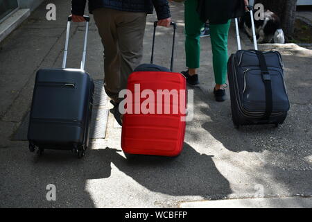 Drei Koffer, Reisetaschen, auf Rädern. Zwei schwarze und eine rote in der Mitte auf dem Gehweg der Straße. Sichtbare Hand von Leuten Koffer ziehen Stockfoto