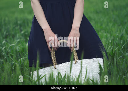 Vorderansicht Nahaufnahme von Frau trug ein schwarzes Kleid und in ihren Händen hält eine weiße Weide-Tasche in einem grünen Feld Weizen Stockfoto