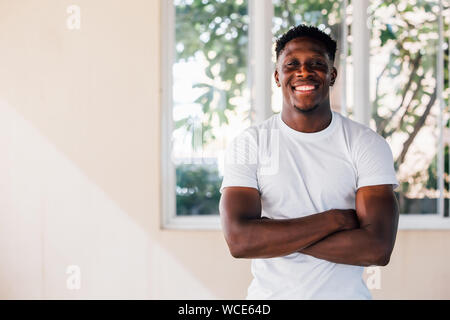 Fröhlich Afrikanische amerikanische Sportler im weißen T-Shirt stand mit verschränkten Armen und lächelnd in die Kamera durch die Fenster in der Turnhalle Stockfoto