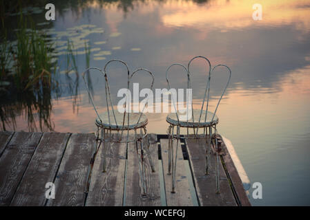Zwei alte Stühle am Rande der Pier vor dem Hintergrund des Sees, der am Morgenhimmel widerspiegelt. Friedlichen Farben Natur Hintergrund. Stockfoto