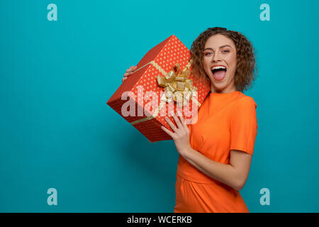 Positive junge Frau in orange Kleid auf Kamera und Schreien während Große in Händen zu halten. Gerne Frau posiert und Lachen auf blauem Hintergrund isoliert. Konzept der Überraschung. Stockfoto