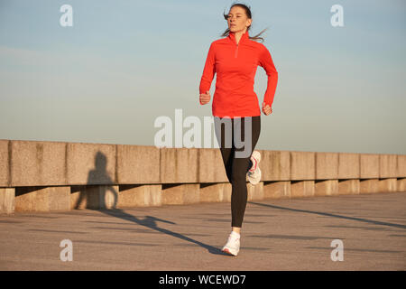 Ein joggingpfad Frau in Rot mit Hemd und schwarze Leggins auf der Straße bei Sonnenaufgang. Auf konkrete Quay. Stockfoto
