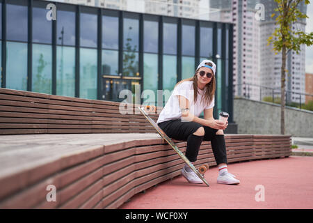 Bild von brunette Sportlerin in Sonnenbrille mit Glas in ihren Händen sitzen auf holzbank in der Nähe von Skateboard auf Sommertag in der Stadt Stockfoto