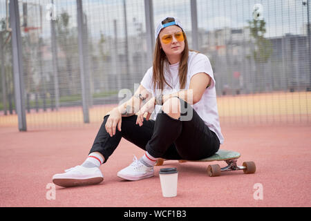 Bild des athletischen brunette Mädchen in Sonnenbrille sitzen auf Skateboard auf dem Spielplatz am Sommertag in der Stadt Stockfoto