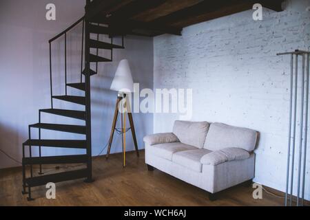 Breites Bild eines weißen Sofas in der Nähe eines Lampenständers In einem Raum mit schwarzer Wendeltreppe Stockfoto