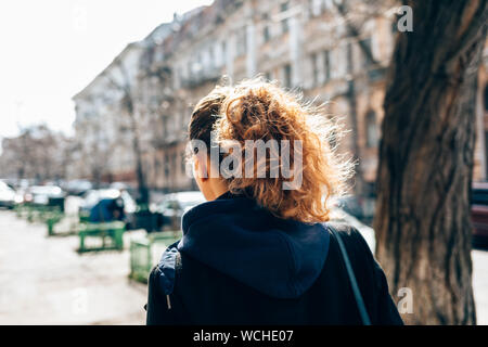 Junge Frau in schwarzen Mantel und mit dem lockigen Haar zu Fuß durch eine alte europäische Stadt Straße neben der Straße und Autos auf Herbst Tag, Ansicht von hinten. Stockfoto