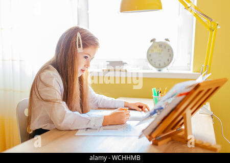 Bildung zu Hause Konzept - süße kleine Mädchen mit langen Haaren Studium oder Abschluss home Arbeit auf einem Tisch mit Stapel Bücher und Arbeitsbuch. Stockfoto