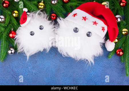 Zwei lustige weißen pelzigen Schnee mann Spielzeug in rot Santa Claus Hüte grünen Tannenzweigen auf blauem Hintergrund niedlichen kleinen Yeti oder Schneeflocken Weihnachten feiern. Stockfoto