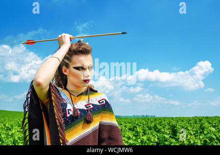 Amazon Mädchen aggressiv Make-up, in einem Poncho, Cosplay im Feld, hält einen Pfeil und Bogen. Heidentum, Feminismus, der Unabhängigkeit, der Stamm Stockfoto
