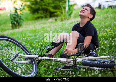 Konzept Fahrrad Unfall, Junge mit Schmerzen in den Kniegelenken nach dem Radfahren mit dem Fahrrad im Park. Knöchel schmerzt nach Fahrrad fallen. Kid Schädigung Stockfoto