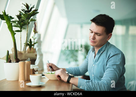 Jungen gutaussehenden Mann sieht auf seine Armbanduhr beim Essen Salat und Kaffee trinken während der Pause im Cafe warten auf seine Patner zu kommen. Stockfoto