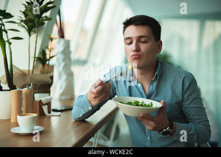 Junge gut aussehender Geschäftsmann setzt Salat in den Mund und trinkt Kaffee zum Mittagessen während der Pause im Cafe in der Nähe von Büro hungrig aussieht. Stockfoto