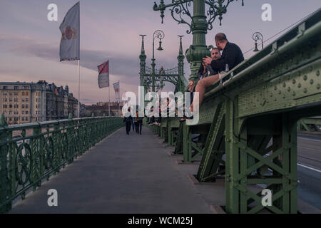 Menschen auf ein Geländer der Brücke in Budapest sitzen und genießen den Sonnenuntergang über der Donau. Stockfoto