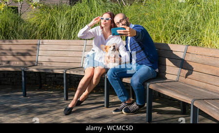 Das glückliche Paar sitzt auf der Bank im Park mit kleinen Hund und Selfie portrait tun. Cute Corgi Pembroke Welpen auf seinen Besitzer. Konzepte Stockfoto