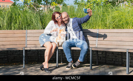 Das glückliche Paar sitzt auf der Bank im Park mit kleinen Hund und Selfie portrait tun. Cute Corgi Pembroke Welpen auf seinen Besitzer. Konzepte Stockfoto