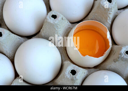 Weißes huhn eier sind frisch, in ökologischen Verpackungen aus Pappe gestapelt. Eines der Eier ist gebrochen und das Eigelb, sichtbar ist. Essen Hintergrund. Close-up. Stockfoto