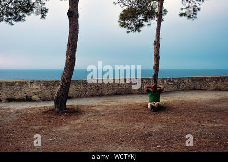 Mann mit einem Pferd Kopf Maske entspannt unter einem italienischen Stein sitzend Kiefer mit dem Meer im Hintergrund Stockfoto