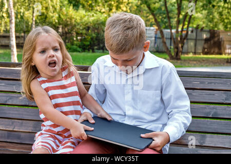 Konzept Gadget kämpfen. Ein Mädchen nimmt einen Laptop von einem Jungen auf einer Bank in einem Park. Stockfoto