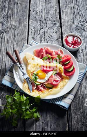 Crêpes mit Spiegelei, Jamon, Scheiben, frischen Tomaten und Gurken Füllungen auf eine weisse Platte auf einem alten Holztisch, vertikale Ansicht von oben Stockfoto