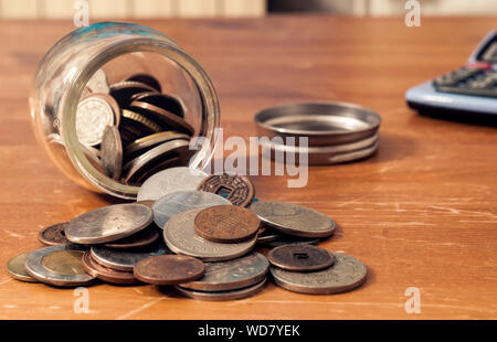 Münze jar umgeworfen auf dem Tisch mit Kappe und Rechner im Hintergrund. Dieses Bild kann dazu verwendet werden, verschiedene Konzepte zu Geld zu vertreten. Stockfoto