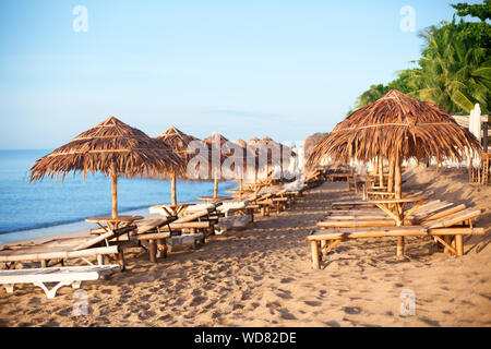 Reihen von leeren Bambus Chaiselongue und strohgedeckten Sonnenschirmen auf einsamen weißen Sandstrand, auf dem blauen Meer und grüne Palmen Hintergrund, niemand, Design Stockfoto