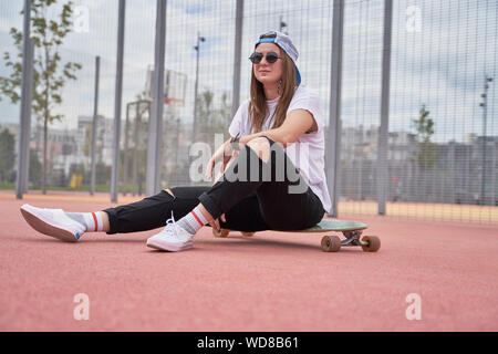 Foto von athletischen brunette Mädchen in Sonnenbrille sitzen auf Skateboard auf dem Spielplatz am Sommertag in der Stadt Stockfoto