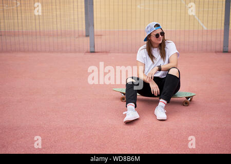 Portrait von athletischen Brünette in Sonnenbrille sitzen auf Skateboard auf dem Spielplatz am Sommertag in der Stadt Stockfoto