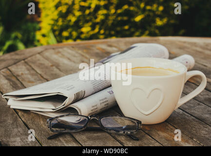 Zeitung, Lesebrillen und eine Tasse Kaffee auf einem Holztisch, im Freien. Stockfoto