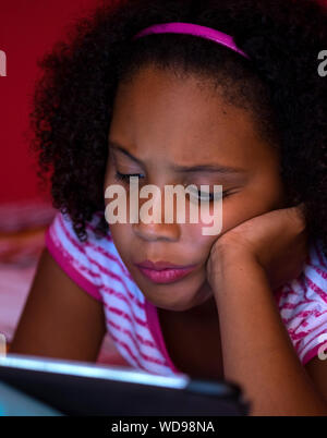 Kind in tiefer Konzentration mit einer schlechten Haltung, spielend auf ihrem Tablet-PC auf ihr Bett, das Tragen eines gestreift rosa und weißen T-Shirt. Stockfoto