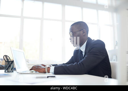 Konzentriert afrikanischen Geschäftsmann in Brillen Tippen auf Laptop Tastatur, während an seinem Arbeitsplatz im Büro zu sitzen Stockfoto