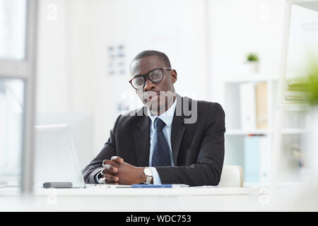 Porträt von Ernst afrikanische Mann Führer in Brillen bei Camera suchen, während Sie im Büro Schreibtisch bei modernen, hellen Büro arbeiten Stockfoto