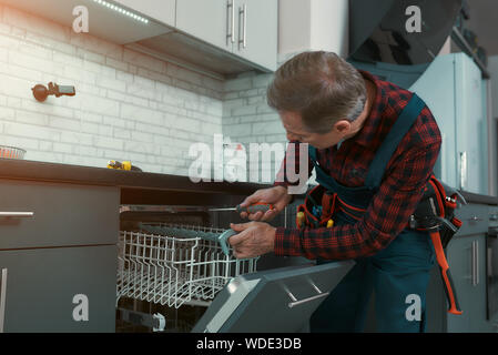 Portrait von reifer Mann tragen tool kit stand in der Küche, mit einem Schraubendreher und Festsetzung der Spülmaschine. Horizontale Schuß Stockfoto