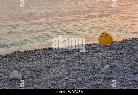 Eine gelbe Kugel auf einem Kieselstrand bei Sonnenuntergang. Kugel mit Smiley. Stockfoto