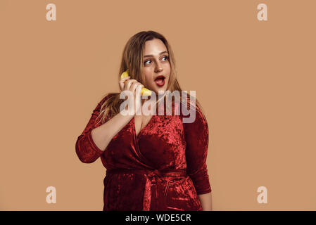 Spielerische. Studio shot Größe lächelnde Frau in Rot velour Kleid holding Banane in der Nähe von ihr Ohr plus und das Gespräch am Telefon. Sorglose Zeit. Übergewichtige Frauen. Xxl-Modell Stockfoto