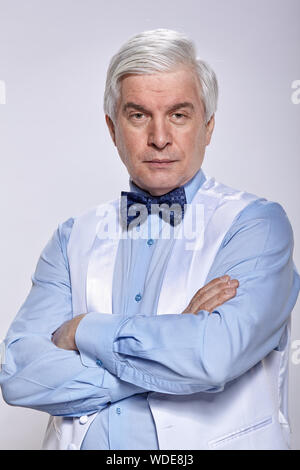 Studio Portrait von älteren Mann mit einem ernsten Gesichtsausdruck auf grauem Hintergrund. Reifer Mann im blauen Hemd mit Weste und Fliege. Ein Blick auf die Kamera Stockfoto