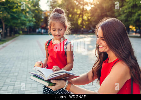 Glückliche Mutter traf ihre Tochter nach dem Unterricht im Freien Grundschule. Frau brachte ihr Kind Bücher und Apple. Familie lesen Buch Stockfoto