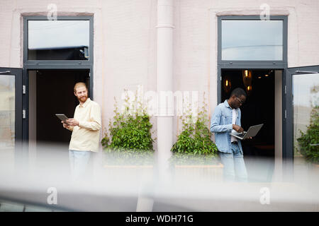 Zwei junge Männer Rücken an Rücken und die Verwendung von Minianwendungen für online arbeiten Sie im Freien in der Nähe des Cafe Stockfoto