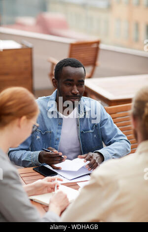 Afrikanische jungen Mann in Freizeitkleidung Notizen in seinem Notizblock beim Sitzen und im Gespräch mit seinen Kollegen an Treffen im Café im Freien Stockfoto