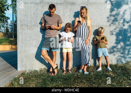 Bild des blonden Frau und Mann und Kinder mit Handys in den Händen stehend auf Betonwand auf der Straße im Sommer Tag Stockfoto