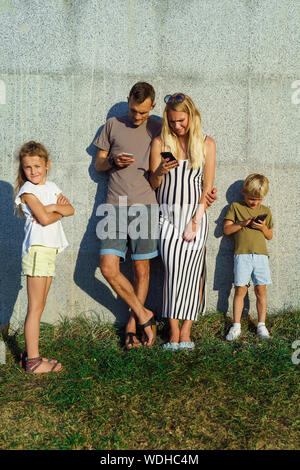 Bild von Frau, Mann und Sohn mit Telefonen in den Händen und Töchter neben Beton Wand draußen auf Sommer Tag Stockfoto