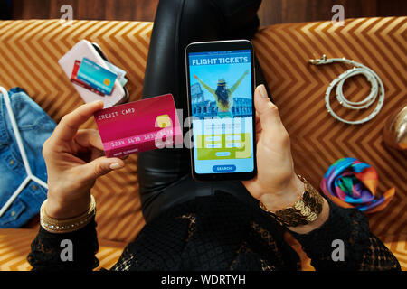 Elegante Hausfrau mit Kreditkarte Kauf von Flugtickets online auf einem Smartphone beim Sitzen auf der Couch im Wohnzimmer. Stockfoto