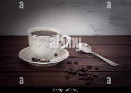 Kaffee in eine Tasse mit Untertasse auf einem Holztisch. Kaffeebohnen sind auf den Karten verstreut und einem silbernen Löffel liegt. Unscharfer Hintergrund weiß mit Farbe Stockfoto