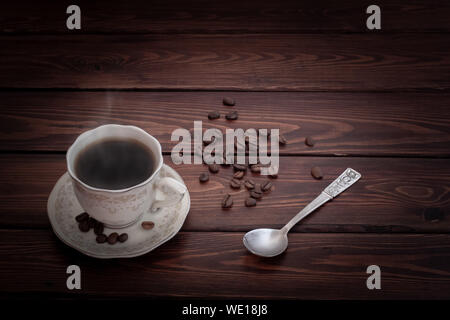 Heiß dampfenden Kaffee in eine Tasse und Untertasse auf einem Tisch mit Kaffeebohnen bestreut. Einen silbernen Löffel liegt in der Nähe. Braun Boards. Für Text platzieren. Stockfoto