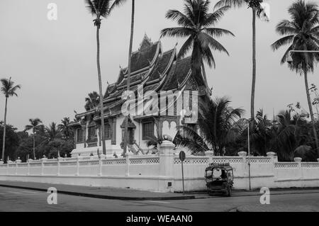 Luang Prabang Royal Palace Museum und Hor Prabang Temple Hall unter der Kokospalme mit Tuk Tuk Park an der Wand Seite - Schwarz und Weiß Stockfoto